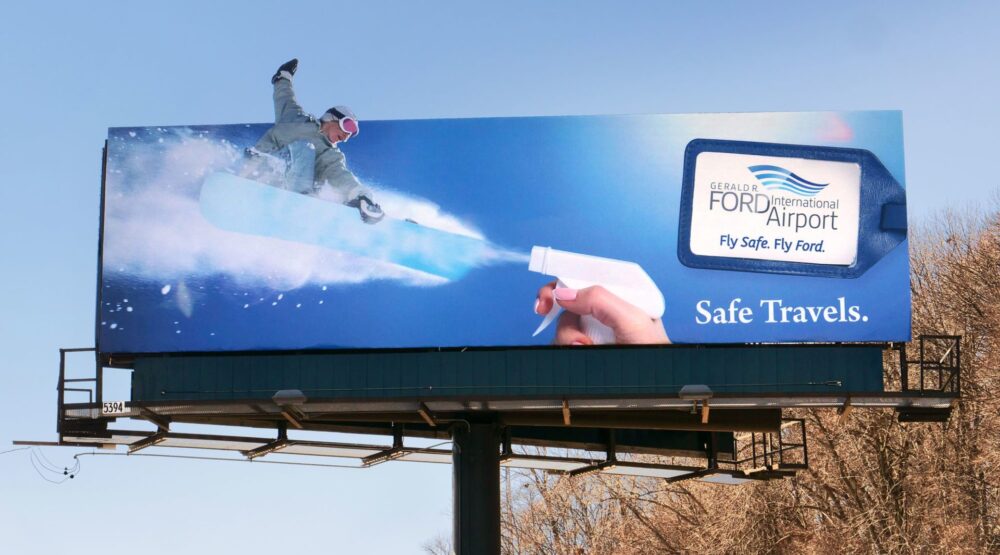 Billboard of someone holding a spray bottle and a snowboarder “riding” the spray, and the headline,”Safe travels.”