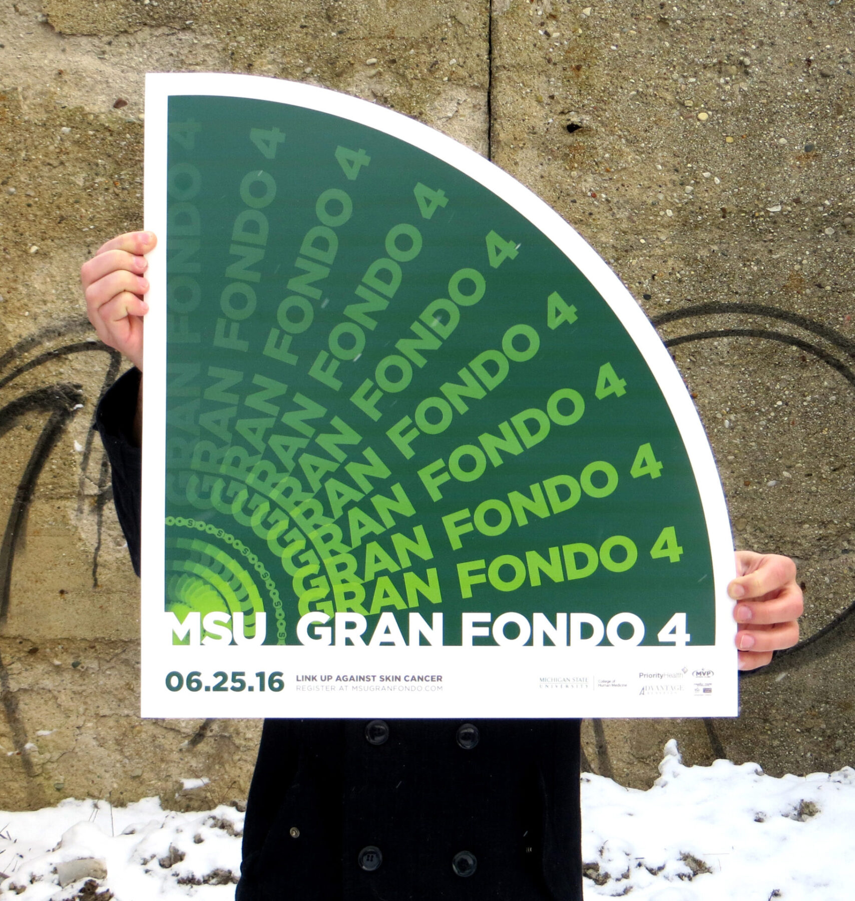 Person holding the MSU Gran Fondo 4 event promotional poster in front of a concrete wall.