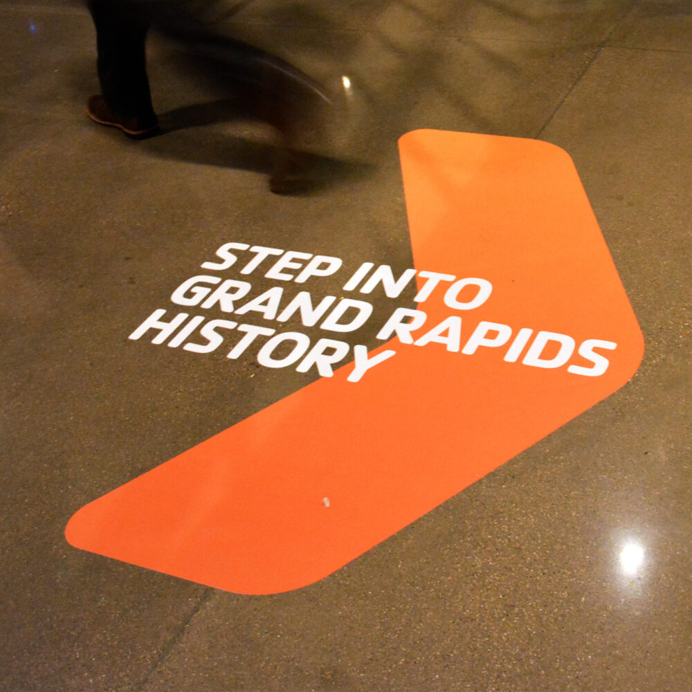 Floor graphic designed for the YMCA featuring a bold orange arrow and the headline, “Step into Grand Rapids history.”