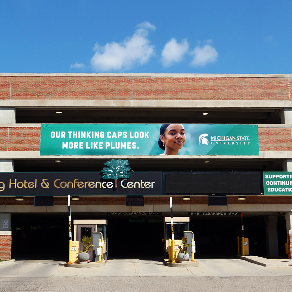 Parking garage banner featuring an image of a student with the headline, “Our thinking caps look more like plumes.”